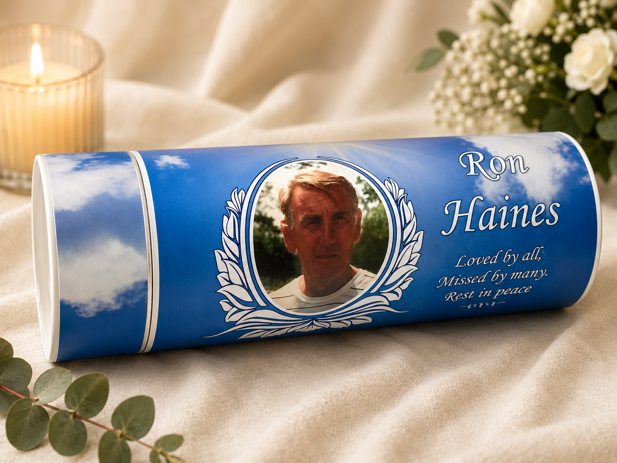 A personalized blue cylindrical urn with a printed portrait of a man, white decorative text reading 'Ron Haines' and a memorial message, styled on beige fabric with a lit candle, flowers, and greenery.