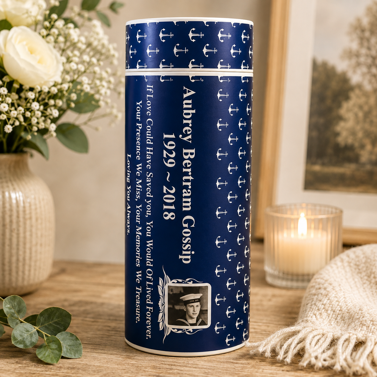A navy blue cylindrical keepsake or memorial box with white anchor patterns and personalized text reading 'Aubrey Bertram Gossip 1929 ~ 2018,' shown on a wooden surface with a flower vase, framed print, and lit candle in the background.