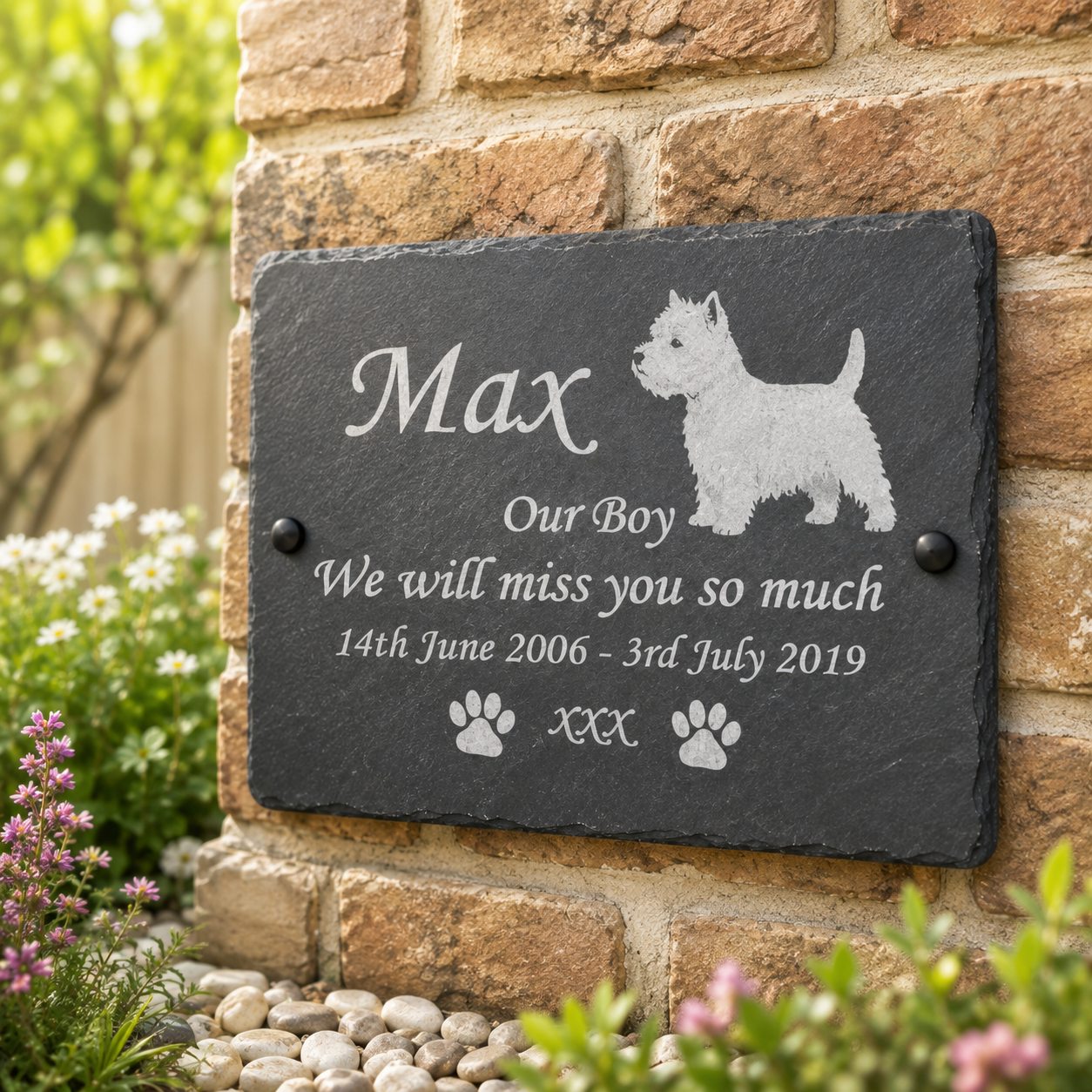 Personalized black slate pet memorial plaque engraved with the name 'Max', a West Highland terrier silhouette, and remembrance text, mounted on a brick wall outdoors with flowers and greenery around it.