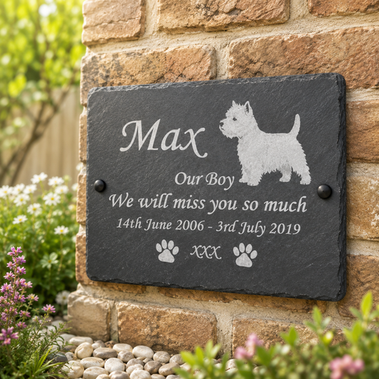 Personalized black slate pet memorial plaque engraved with the name 'Max', a West Highland terrier silhouette, and remembrance text, mounted on a brick wall outdoors with flowers and greenery around it.