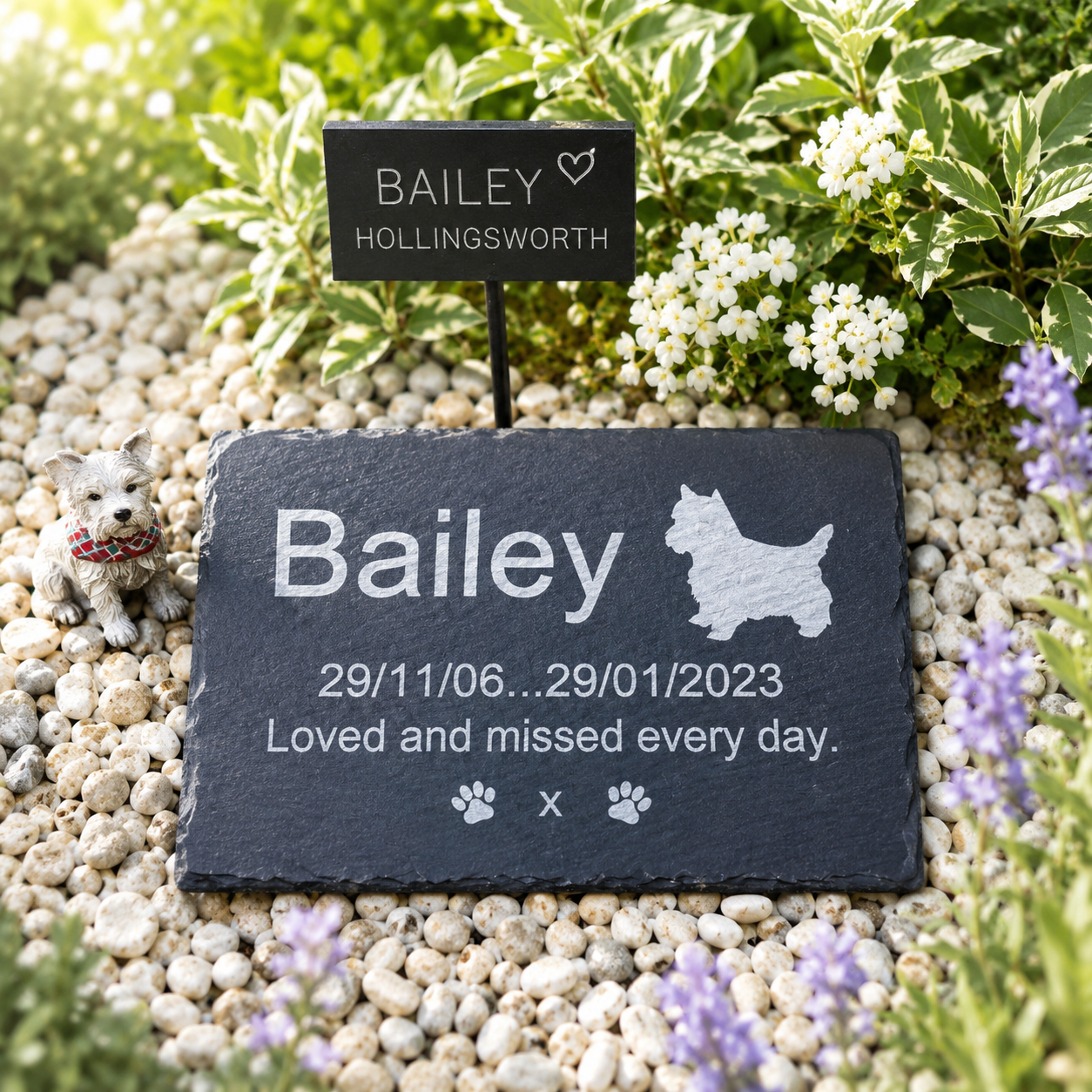 Personalized pet memorial garden plaque in a real garden setting, surrounded by gravel, greenery, and small flowers. The main product is a dark slate stone plaque engraved with the name Bailey, a silhouette of a Scottish Terrier, dates, and a memorial message.