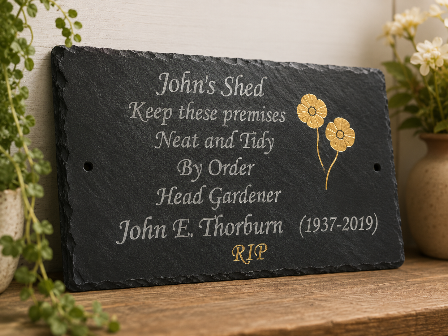 Personalized black slate memorial plaque with engraved white text and gold floral accents, displayed on a wooden surface with potted plants in the background.