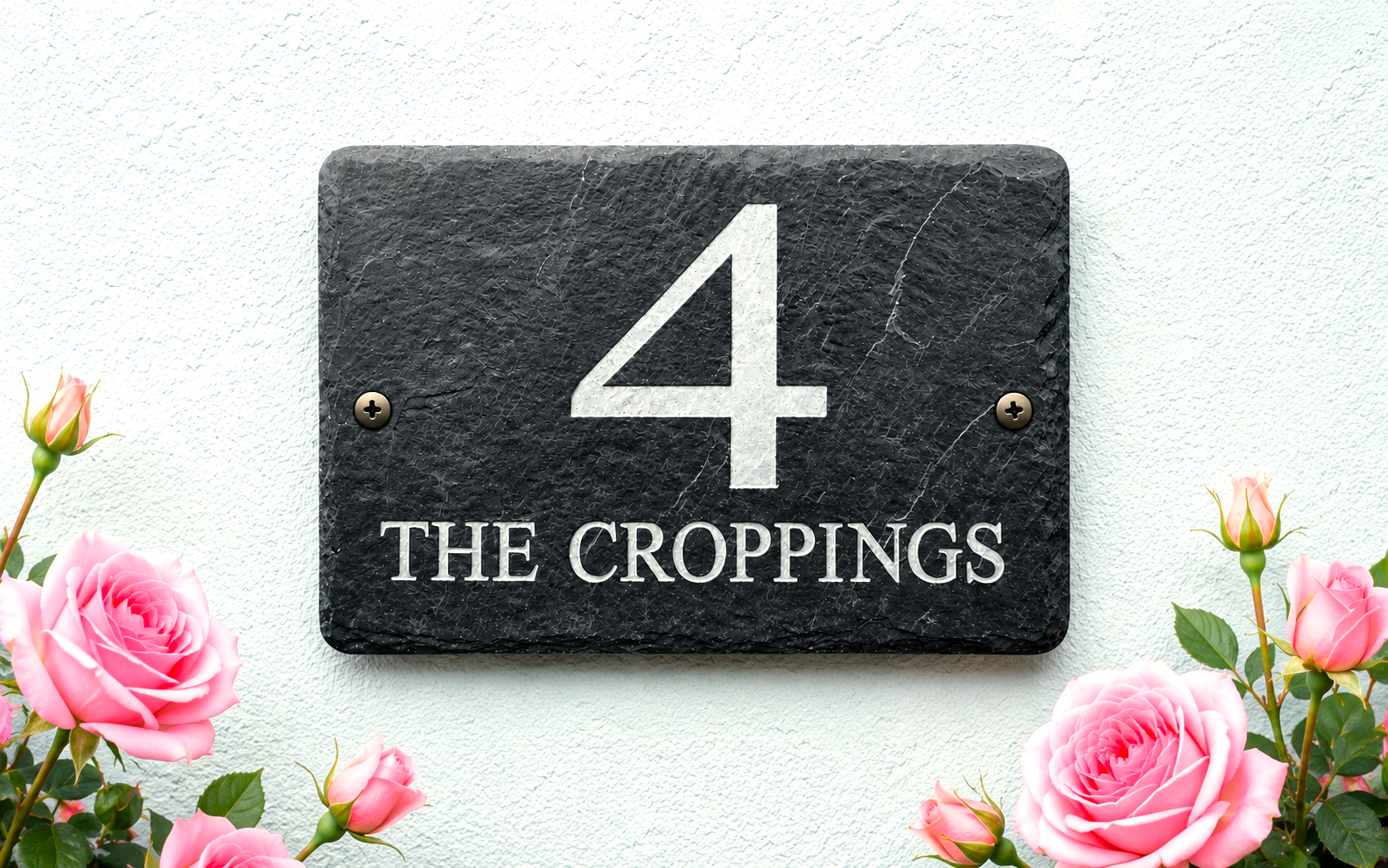 A rectangular black slate house number plaque reading '4 THE CROPPINGS' in white lettering, mounted on a textured white wall and framed by pink roses at the bottom corners.