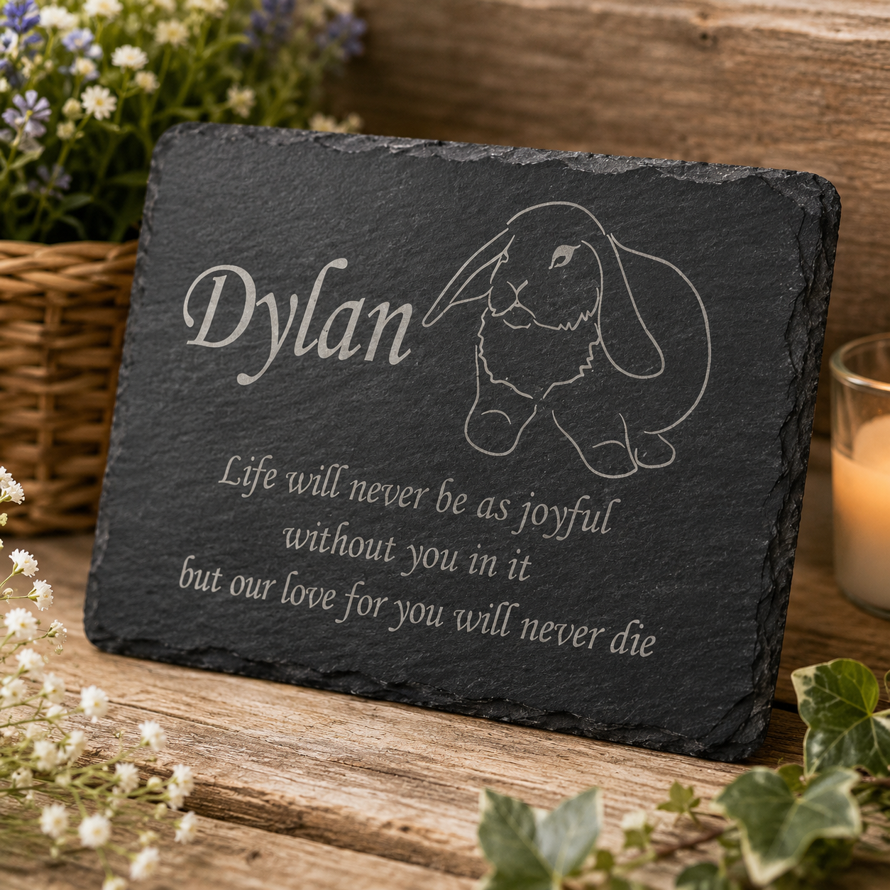Personalized black slate memorial plaque engraved with the name 'Dylan', a bunny illustration, and a remembrance quote, displayed on a rustic wooden surface with flowers and a candle in the background.