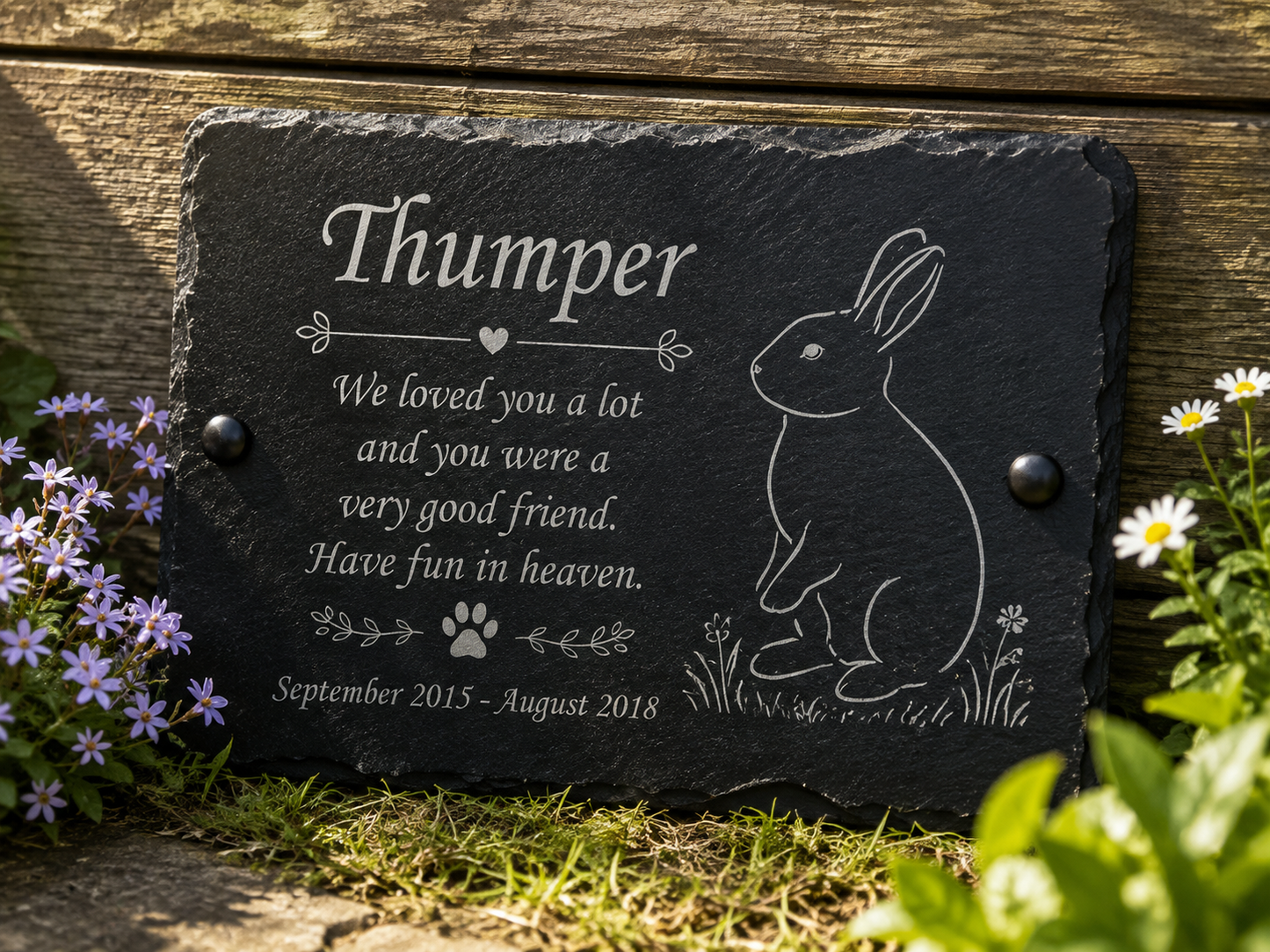 Personalized black slate pet memorial plaque engraved with the name 'Thumper', a rabbit illustration, and remembrance text, displayed outdoors against a wooden fence with flowers around it.