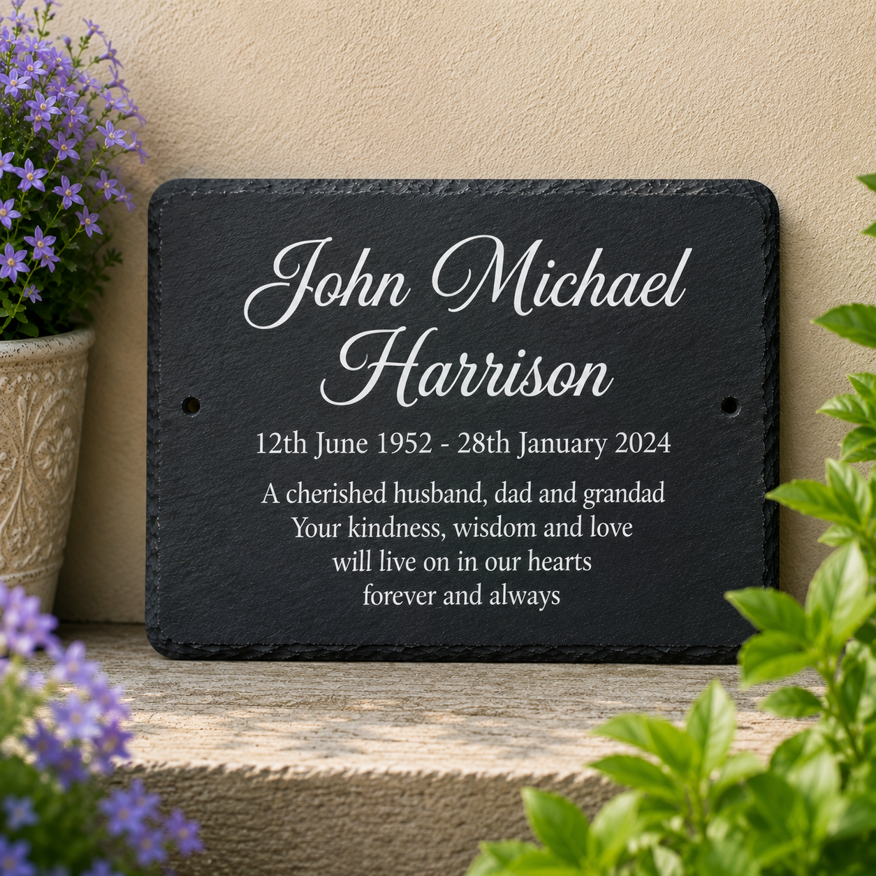 Personalized black slate memorial plaque with white engraved text, displayed on a stone ledge with potted flowers and greenery against a beige wall.