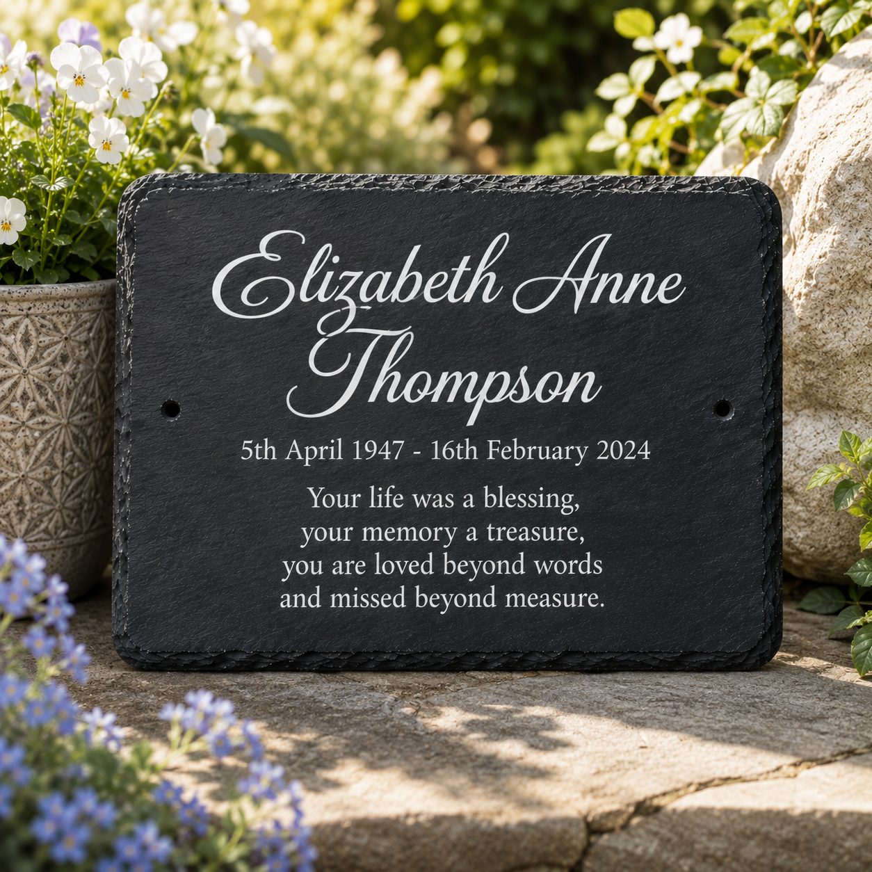 Personalized black slate memorial plaque with white engraved text reading 'Elizabeth Anne Thompson' and a remembrance message, displayed outdoors on a stone surface with flowers and rocks in the background.