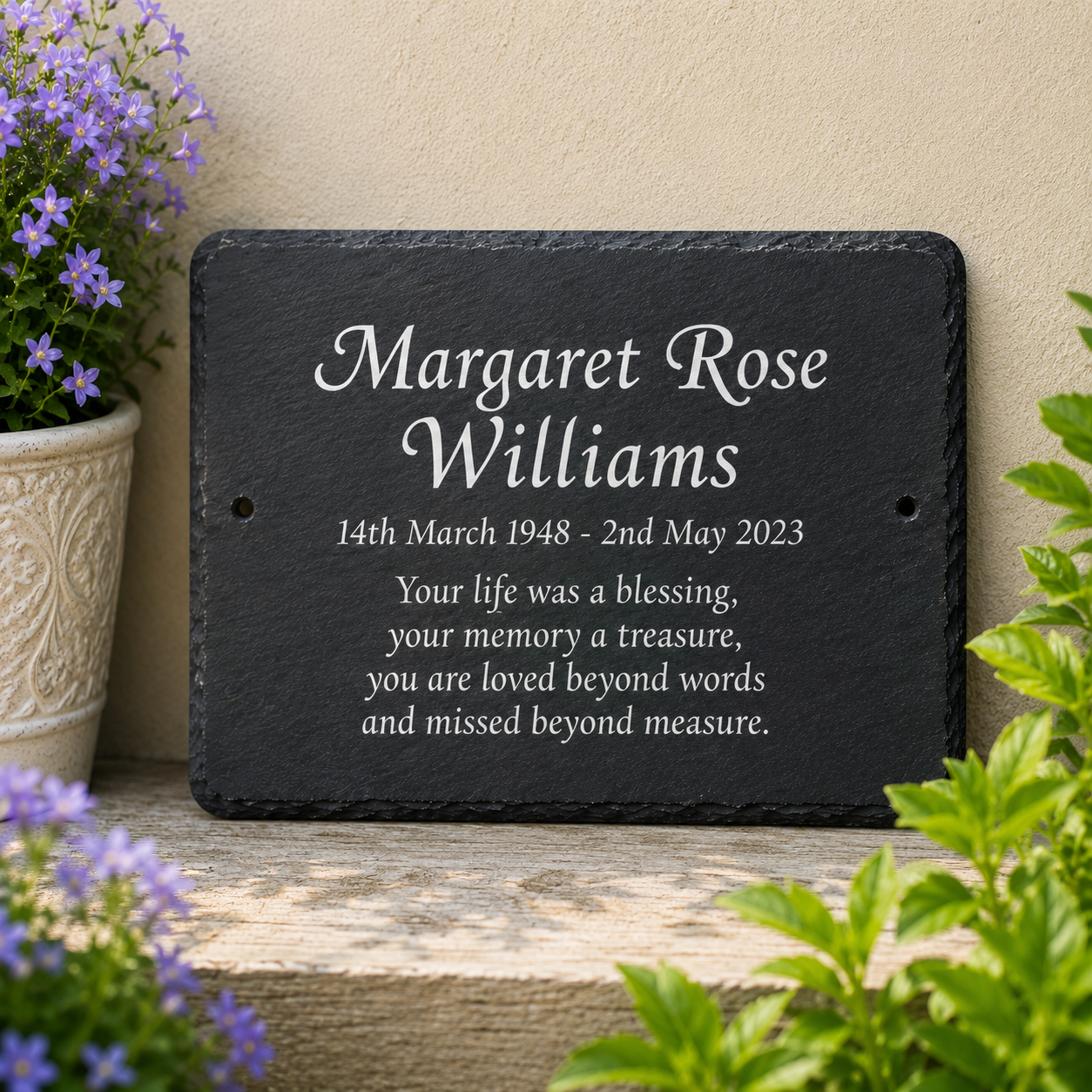 Personalized black slate memorial plaque with white engraved text, displayed on a wooden ledge against a beige wall with potted plants nearby.