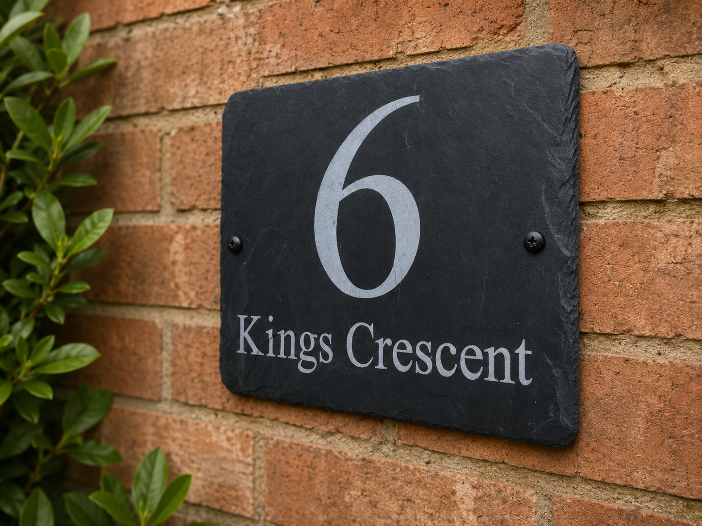 A rectangular black slate house number plaque reading '6 Kings Crescent' mounted on a red brick wall with green leaves visible on the left side.