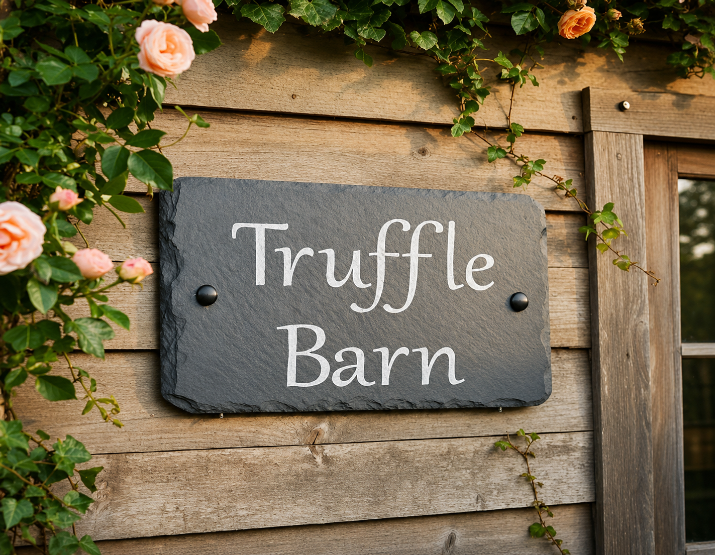 A dark slate sign reading 'Truffle Barn' in white script, mounted on a rustic wooden wall with climbing green vines and pink roses around it.