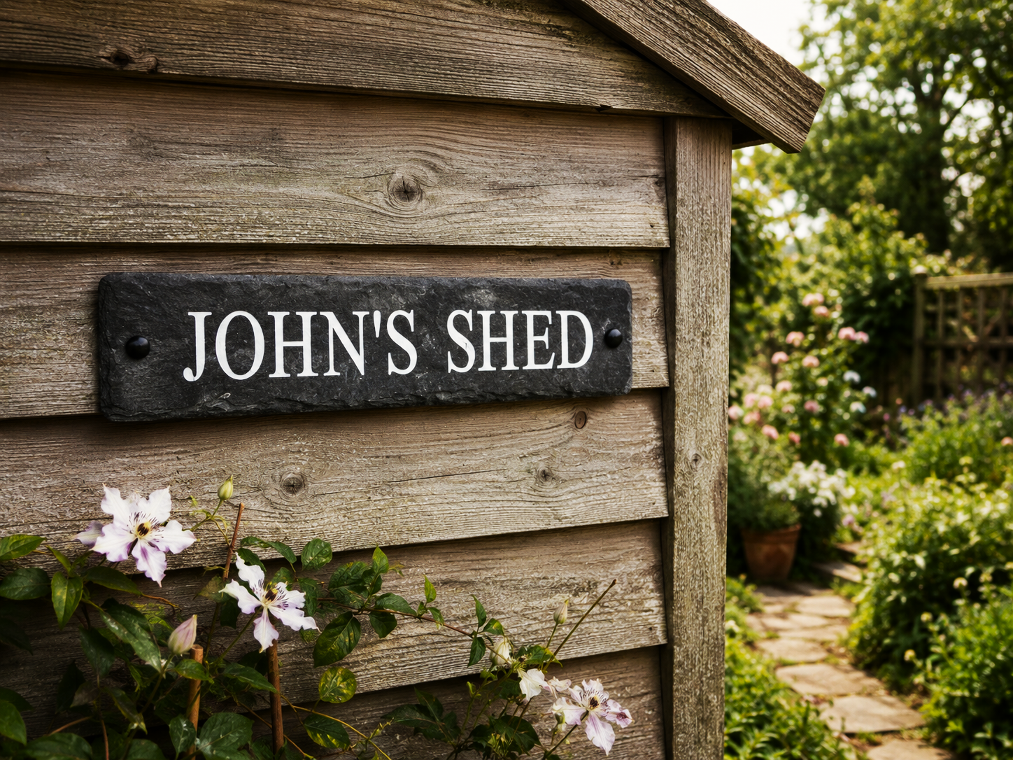 A rustic black stone sign reading 'JOHN'S SHED' mounted on weathered wooden siding, with greenery and small white flowers in the foreground and a garden path in the background.