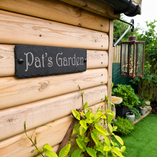 A rectangular black slate garden sign reading 'Pat's Garden' in white script, mounted on a light wood log wall in a garden setting with green plants and grass.