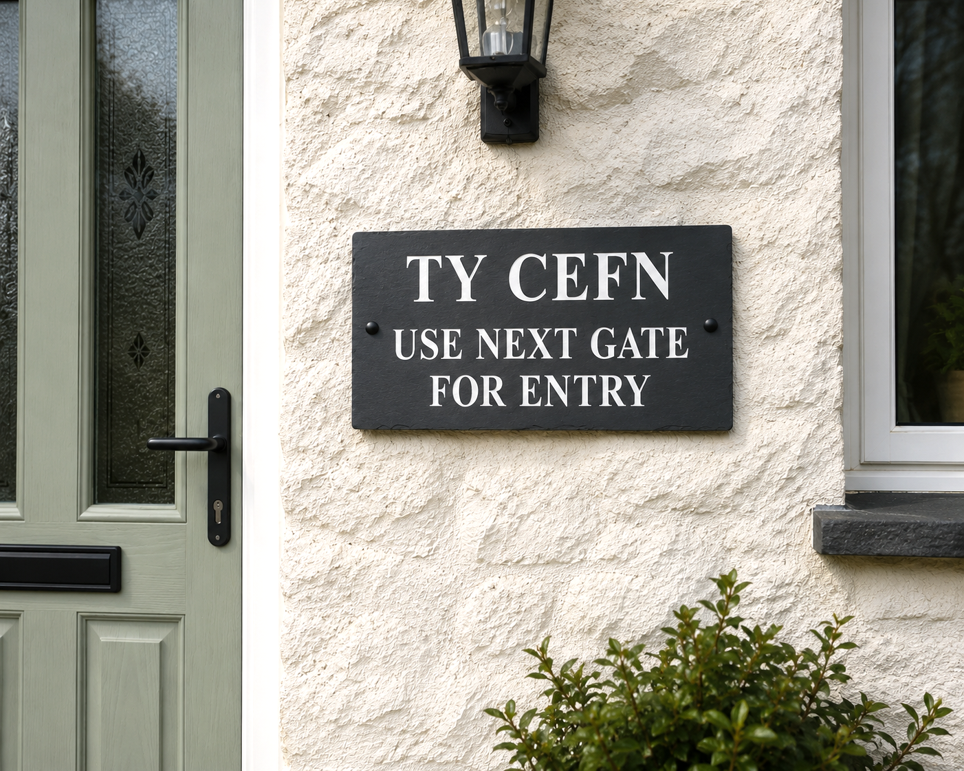 A rectangular black metal sign with white text reading 'TY CEFN USE NEXT GATE FOR ENTRY' mounted on a textured cream stucco wall next to a green front door and beneath a black wall lantern.
