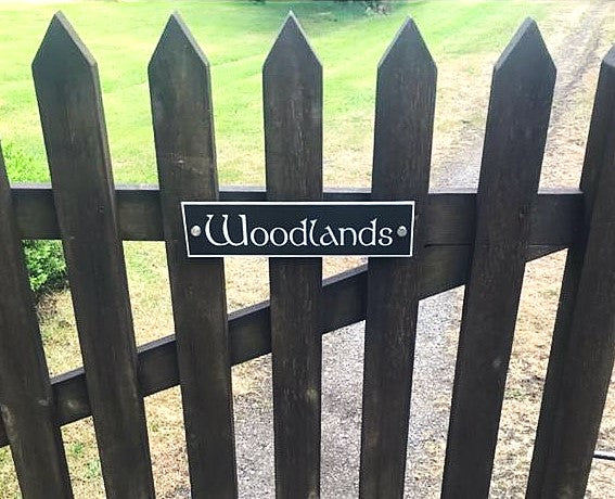 A dark brown wooden picket fence with a central sign that reads 'Woodlands' in white script, set against a background of green grass and a gravel path.