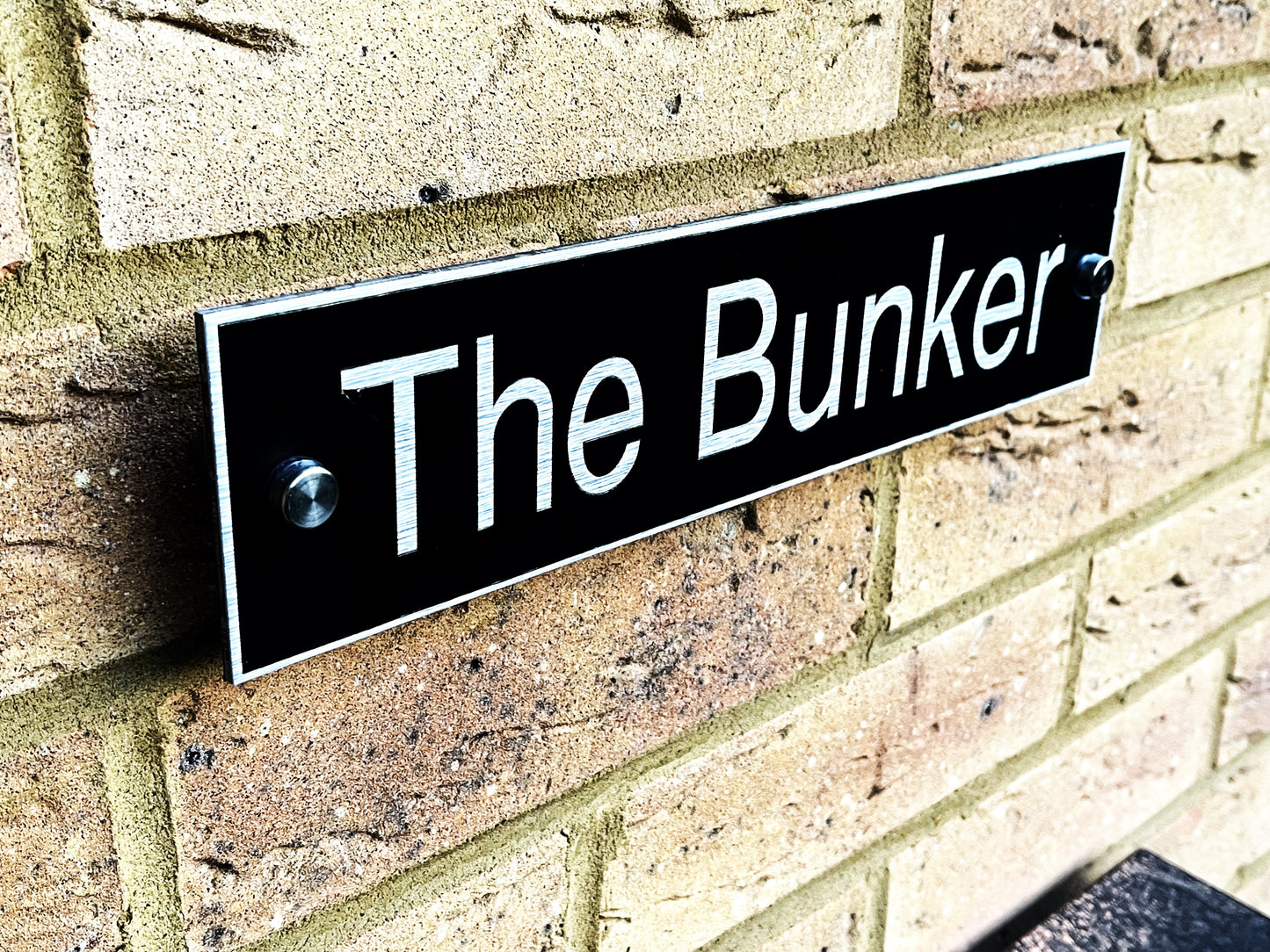 A black rectangular metal sign with white text reading 'The Bunker' mounted on a tan brick wall.