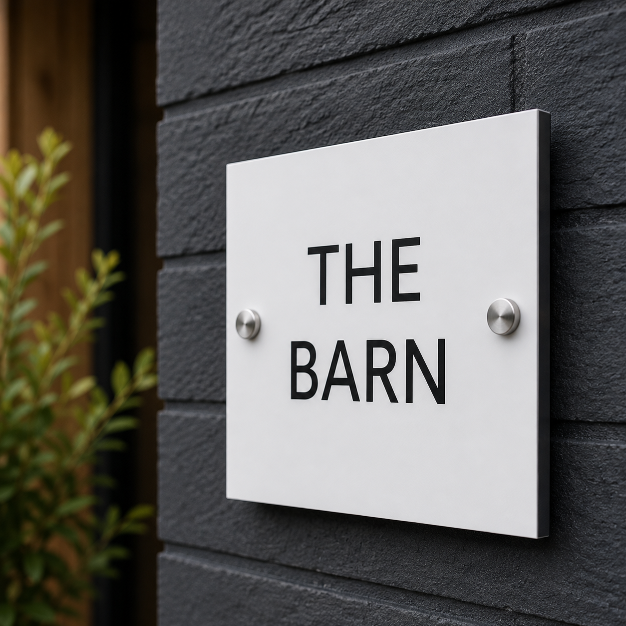 A modern white square wall sign with the words 'THE BARN' in bold black letters, mounted on a dark gray brick wall with two silver screws.