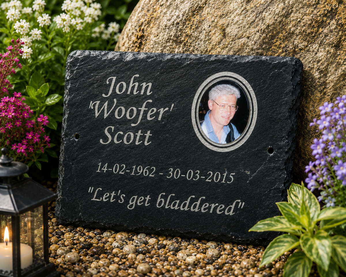 Personalized black slate grave marker with engraved white text, a circular photo portrait of the deceased, and the humorous epitaph "Let's get bladdered," displayed outdoors on gravel beside flowers and a lantern.