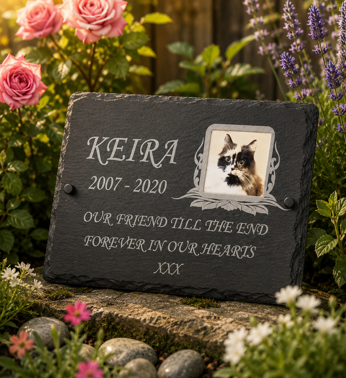 A personalized black slate pet memorial plaque with a photo of a cat and engraved text reading 'KEIRA 2007-2020 OUR FRIEND TILL THE END FOREVER IN OUR HEARTS XXX', displayed outdoors in a garden setting.