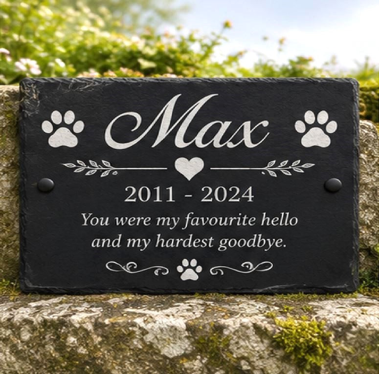 A personalized black slate pet memorial plaque engraved with the name 'Max', paw prints, a heart, and the text '2011 - 2024 You were my favourite hello and my hardest goodbye.' The plaque is mounted on a stone wall with blurred green foliage in the background.