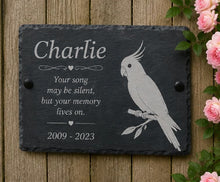 Memorial slate plaque with engraved parrot design and text on a wooden background with flowers.