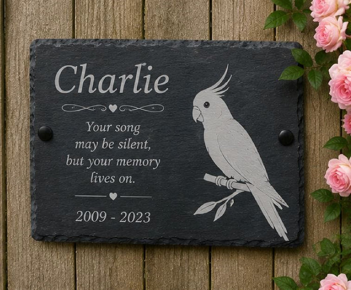 Memorial slate plaque with engraved parrot design and text on a wooden background with flowers.