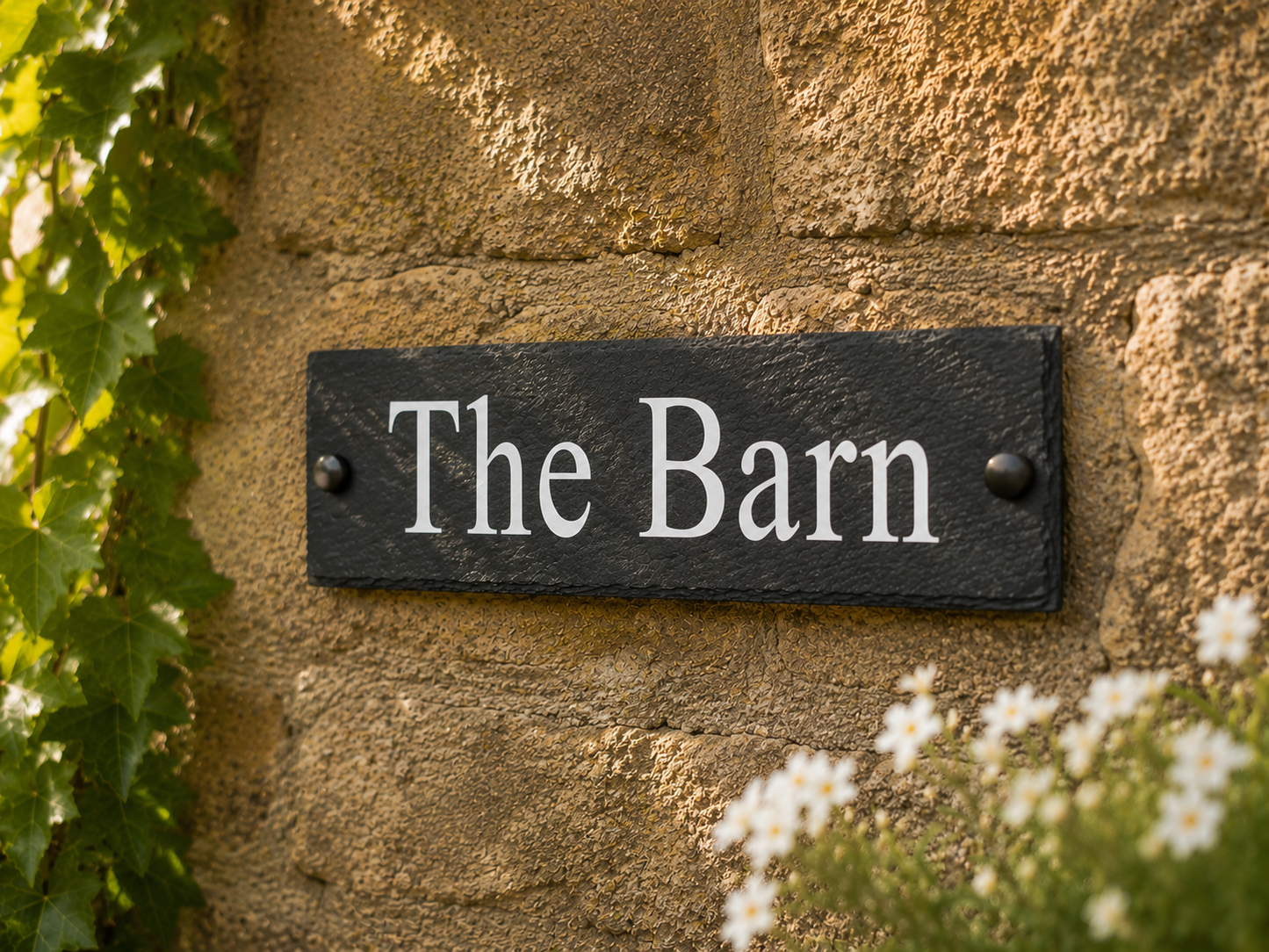 A rectangular black slate sign with white serif text reading 'The Barn,' mounted on a tan stone wall with green leaves and small white flowers around it.