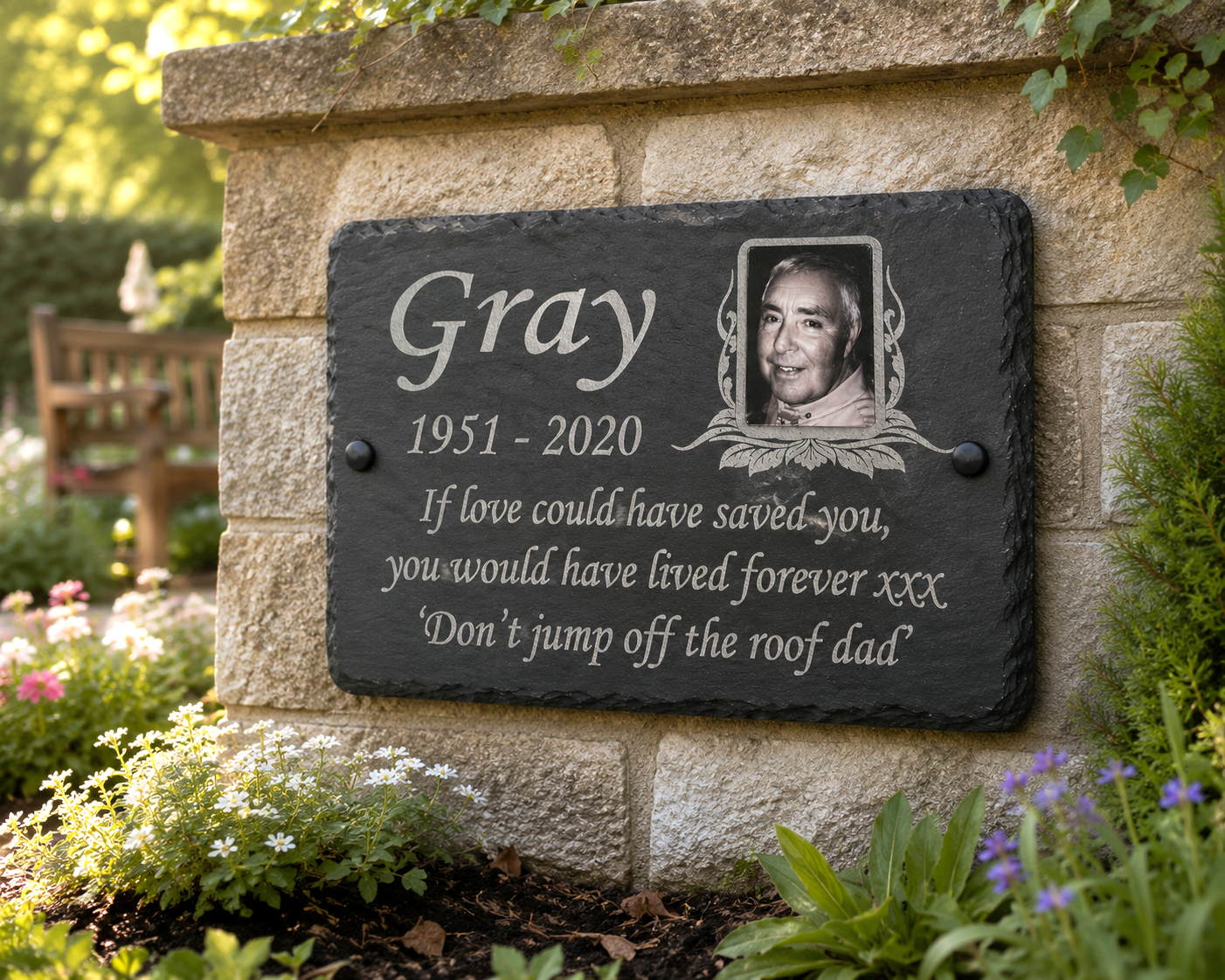A personalized black slate memorial garden plaque mounted on a stone wall, featuring the name 'Gray', a portrait photo, and a remembrance quote, surrounded by greenery and flowers.