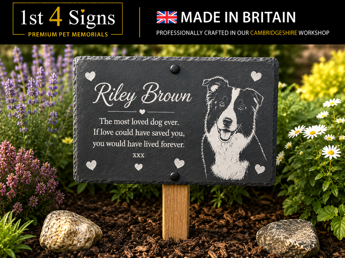 Personalized pet memorial garden plaque on a wooden stake, shown outdoors in mulch with flowers and rocks. The black slate sign has a white engraved-style portrait of a dog and the text 'Riley Brown' and a remembrance message.