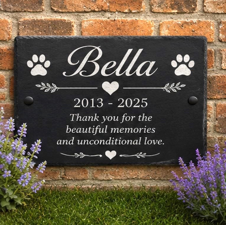 A personalized black slate memorial plaque mounted on a brick wall, featuring the name 'Bella' in script, paw prints, a heart, the years '2013 - 2025', and a remembrance message. Green grass and purple flowers are visible at the base.