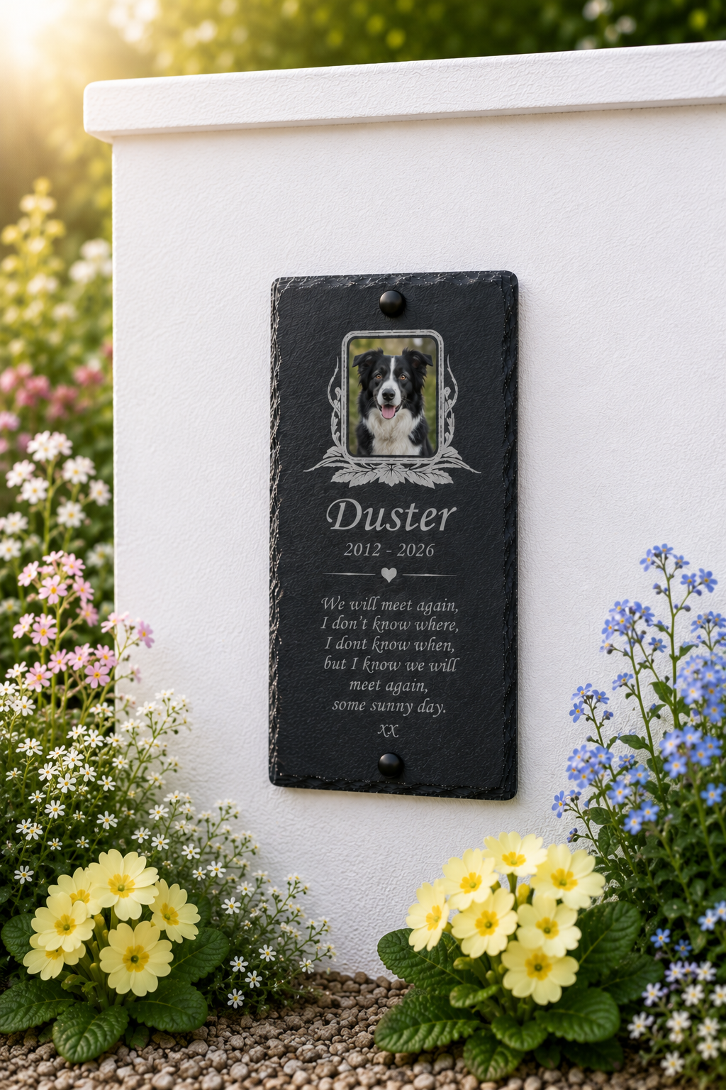 A personalized black slate pet memorial plaque mounted on a white stone wall in a garden setting, featuring a photo of a dog and the name 'Duster' with remembrance text.
