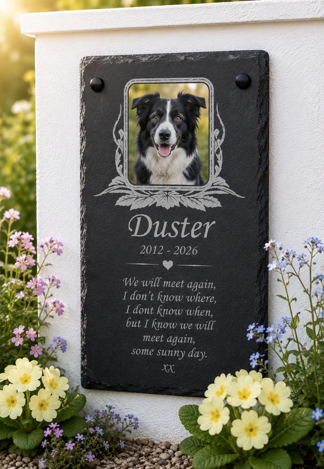 Personalized black slate pet memorial plaque with a photo of a black and white dog, engraved with the name 'Duster' and a remembrance poem, mounted on a white wall in a garden with flowers.