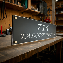 Street sign with address '714 Falcon Mews' on a wooden surface in a workshop setting