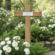 Wooden cross with a plaque in a garden setting with white flowers and greenery.