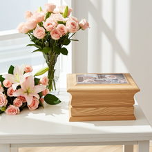 Wooden urn with a photo on top, surrounded by pink flowers on a white surface.