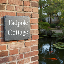 Slate sign reading 'Tadpole Cottage' on a brick wall with a pond and garden in the background.
