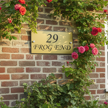 Gold house number sign '29 FROG END' on a brick wall with roses and greenery.