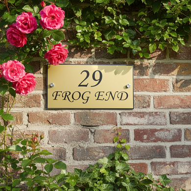 Gold house number sign '29 FROG END' on a brick wall with pink roses.