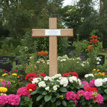 Wooden cross with a plaque in a garden setting with colorful flowers and trees in the background.