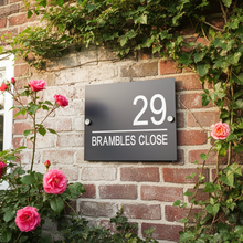 House number sign '29 Brambles Close' on a brick wall with pink roses and greenery.