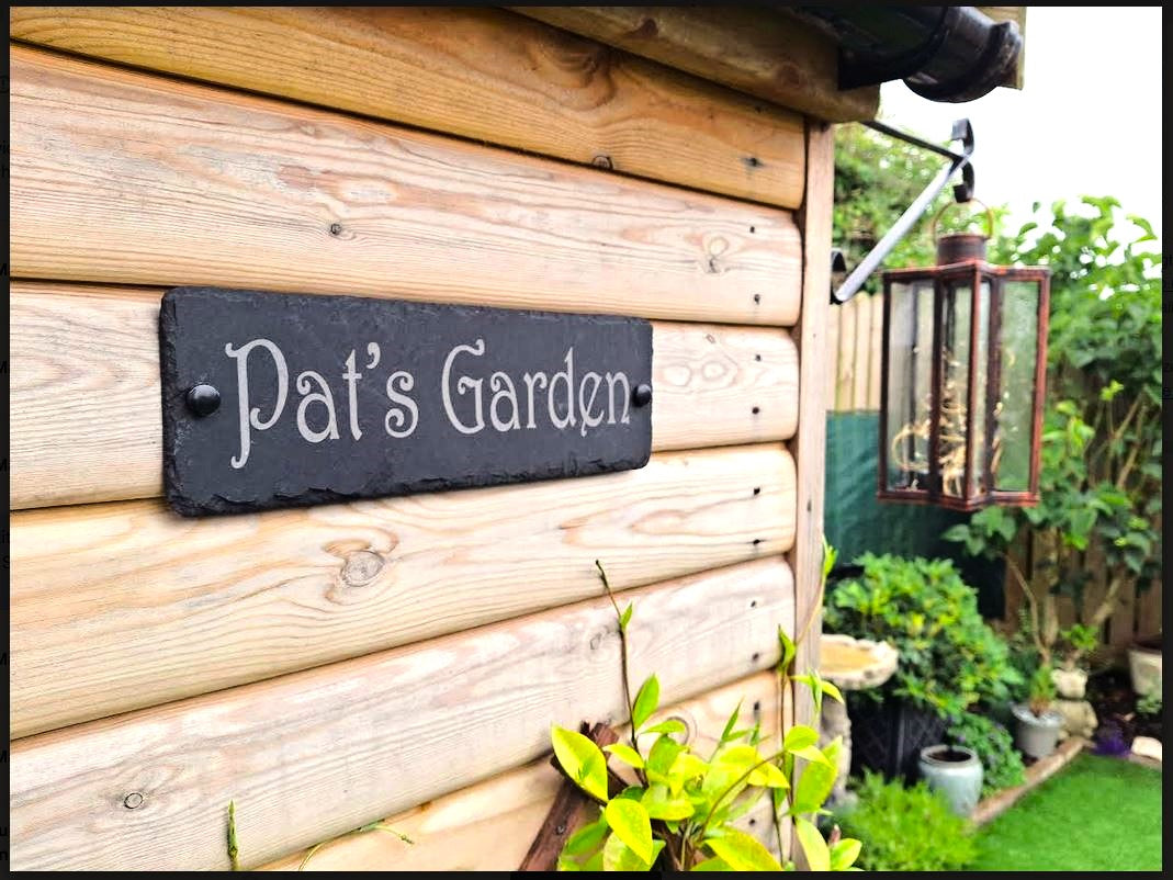 A rectangular black slate garden sign with white script reading 'Pat's Garden' mounted on a light wood log wall in a garden setting.