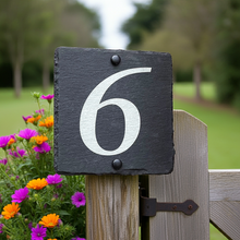 Slate house number '6' on a wooden post with flowers and a blurred green background
