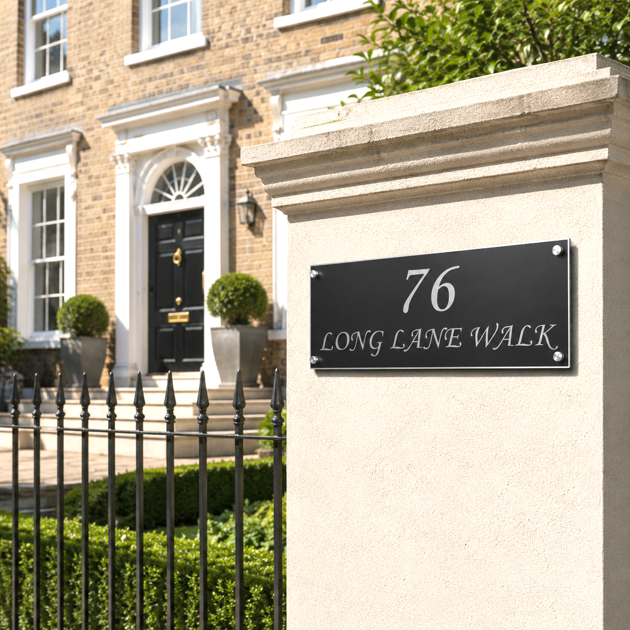 A black rectangular house number plaque with silver lettering reading '76 LONG LANE WALK' mounted on a cream-colored stone pillar in front of a brick house with a black metal fence.