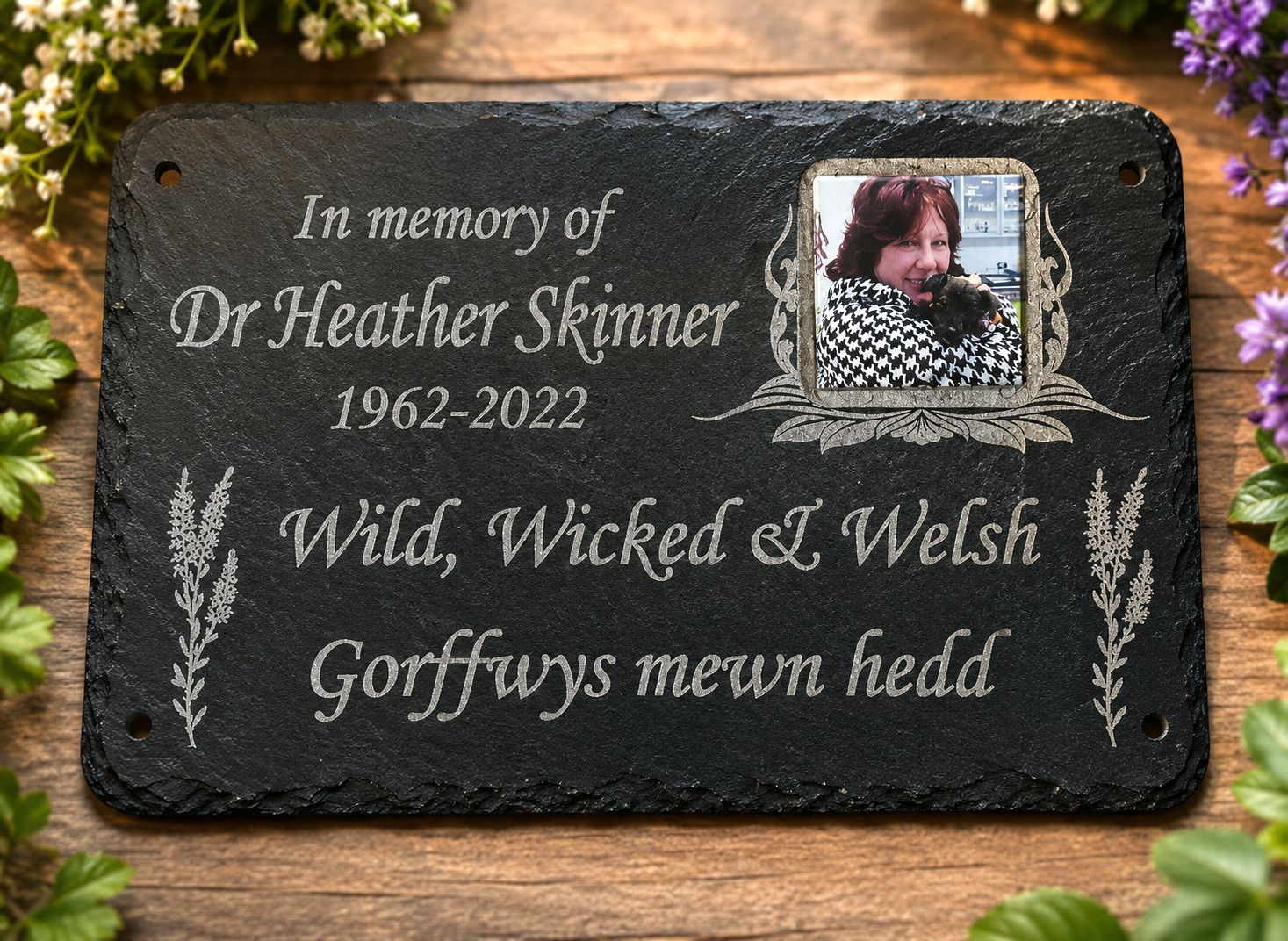 Personalized black slate memorial plaque with engraved white text reading 'In memory of Dr Heather Skinner 1962-2022' and 'Wild, Wicked & Welsh Gorffwys mewn hedd', plus a small inset photo of a woman and decorative botanical flourishes, displayed on a wooden surface with flowers around the edges.