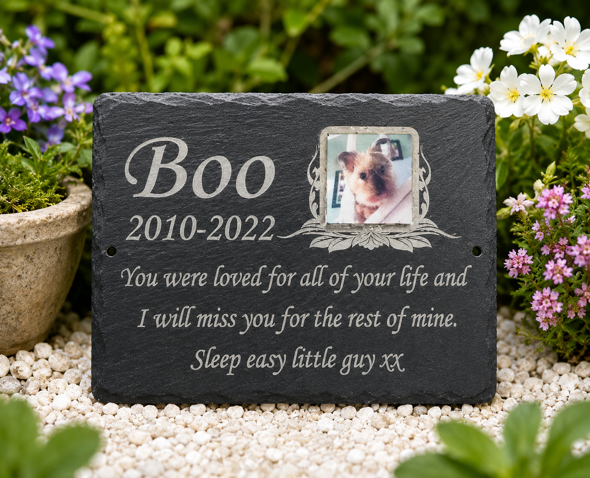 Personalized black slate pet memorial garden stone with engraved text reading 'Boo 2010-2022' and a small photo of a dog, placed on white gravel surrounded by green foliage and flowers.