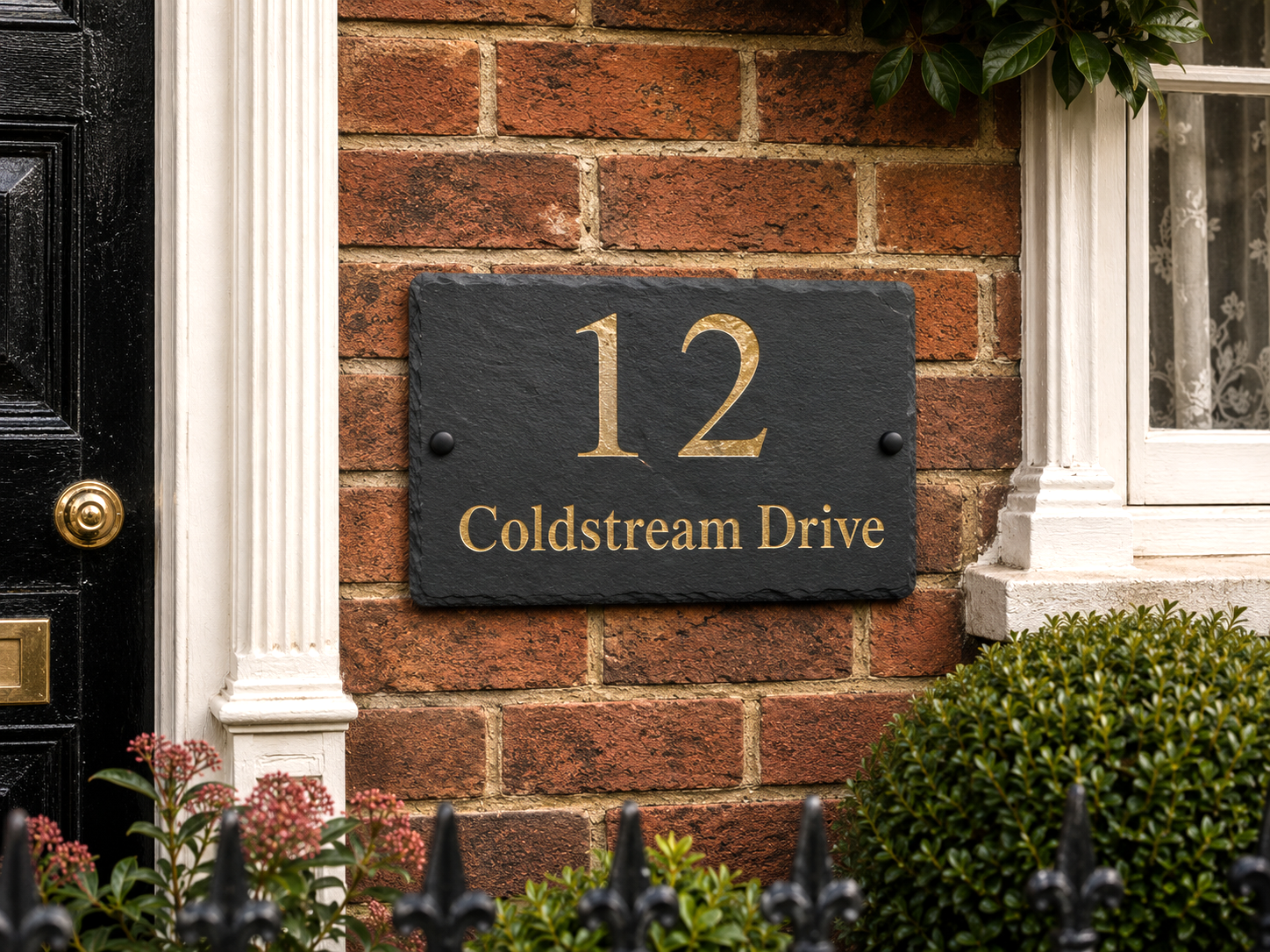 A rectangular black slate house number sign with gold lettering reading '12 Coldstream Drive' mounted on a red brick wall next to a black door and white window trim.