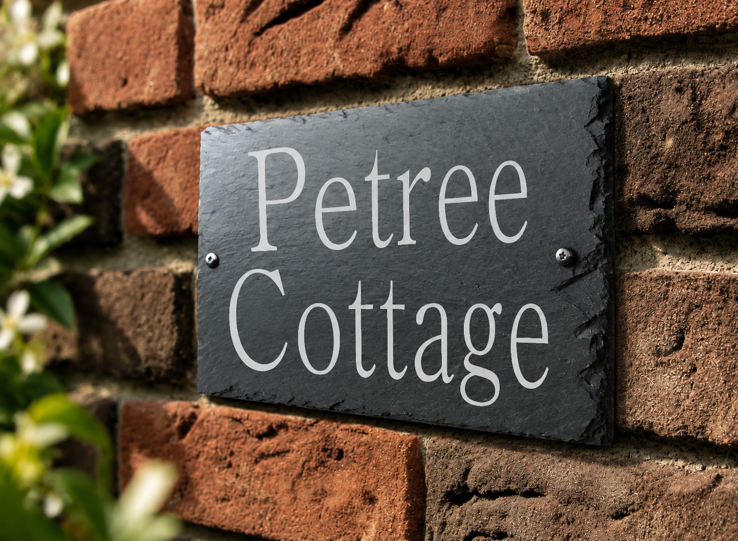 A rectangular black slate house sign with white lettering that reads 'Petree Cottage,' mounted on a red brick wall with blurred greenery at the side.