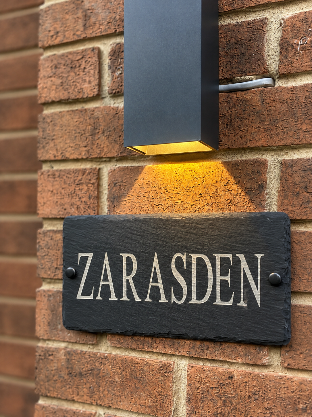 A rectangular black outdoor wall light mounted on a red brick wall above a dark slate house sign reading 'ZARASDEN' in white lettering.