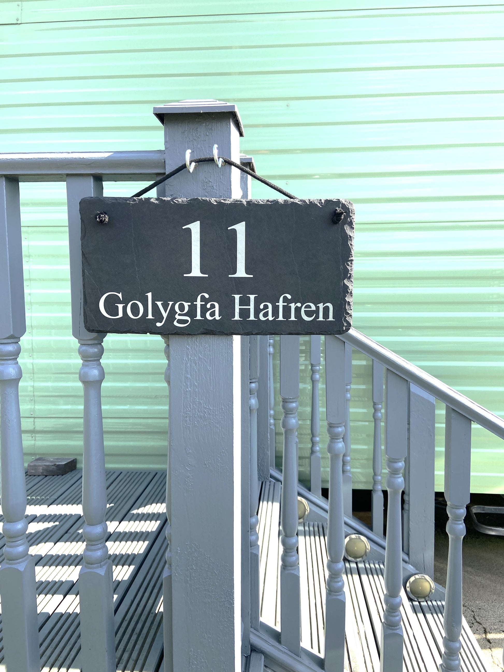 A dark slate house number sign reading '11 Golygfa Hafren' hanging on a gray wooden porch post in front of a mint green corrugated metal wall.