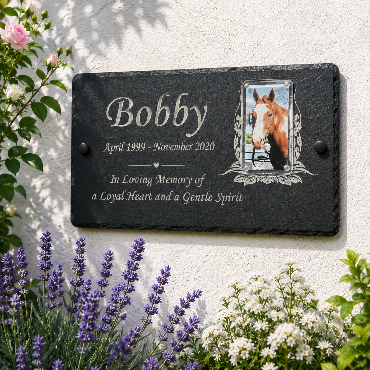 Personalized black slate memorial plaque mounted on a white stucco wall, featuring the name 'Bobby', dates, remembrance text, and a photo frame showing a horse. Purple lavender, white flowers, and green leaves surround the plaque.