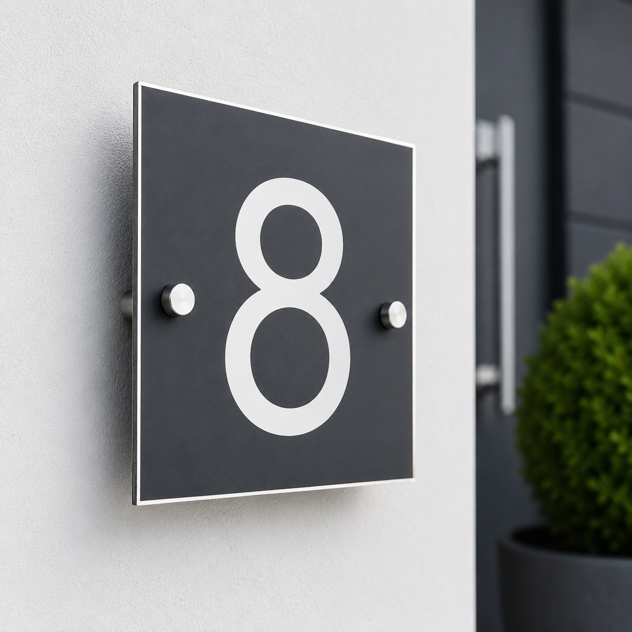 A modern square black house number plaque with a large white numeral '8' mounted on a light gray exterior wall, with a dark door and green shrub blurred in the background.